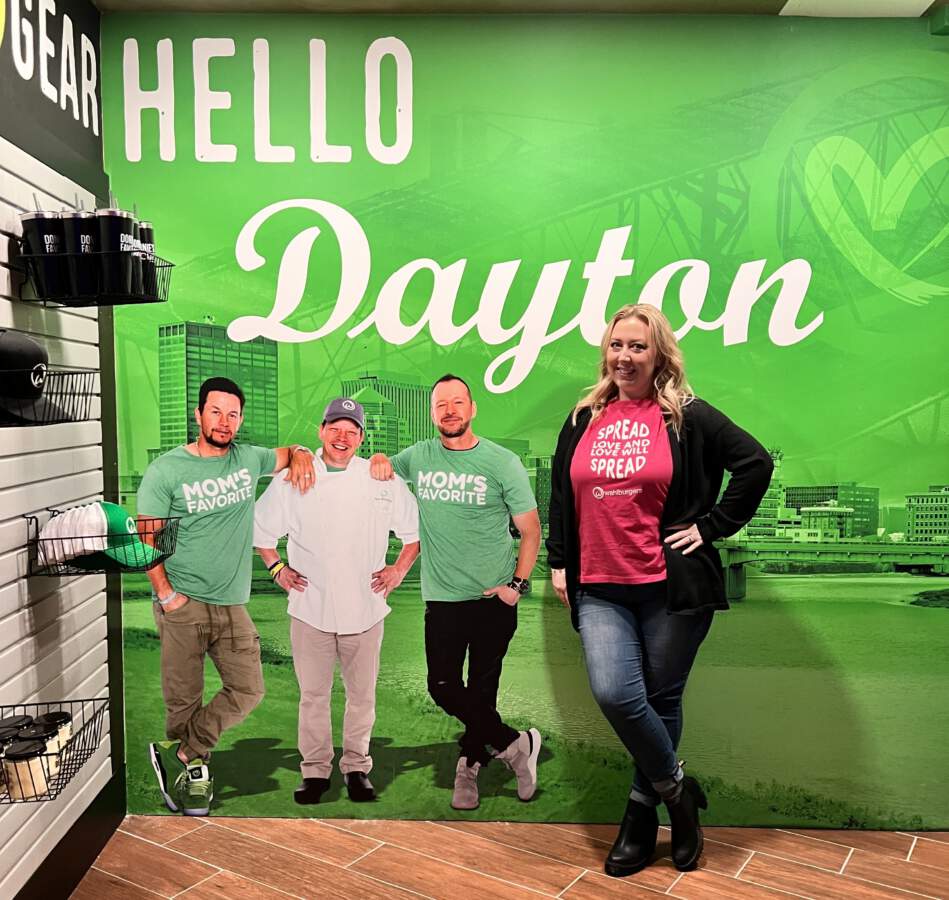 Writer, Nikki Nett, posing in front of green selfie station and mural of Mark, Paul, and Donnie Wahlberg at the Dayton Ohio location of Wahlburgers inside Hollywood Gaming and Racino  